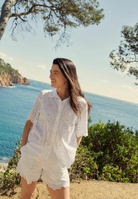 Camicia bianca in pizzo con maniche corte e bordi scallop, abbinata a pantaloni corti bianchi coordinati, sullo sfondo di una costa.
