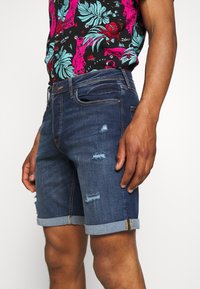 Mies, joka pukeutuu tummansinisiin käännettyihin farkkushortseihin, joissa on kulumisia, ja värikkääseen kukkakuosiselle paidalle, joka on pantu sisään.