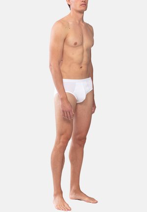 Young adult male standing barefoot wearing white brief-style underwear against a plain light gray background.