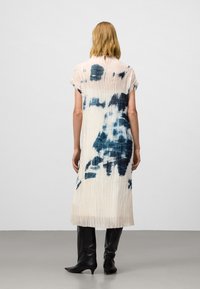 Vrouw met schouderlang blond haar, gekleed in een witte geplooide jurk met blauwe tie-dye patronen en zwarte kniehoge laarzen, staande met haar rug naar ons toegekeerd.