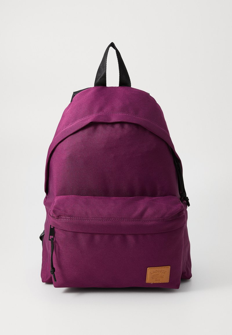 Sac à dos en tissu violet avec une surface texturée, une poche avant et une étiquette en cuir. Il comprend une poignée supérieure et des bretelles noires ajustables.