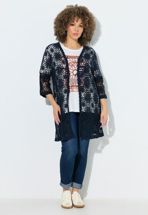 Kvinde med krøllet hår iført en marineblå hæklecardigan, hvid T-shirt med print, mørke opkrogede jeans og hvide sneakers, stående op ad en hvid væg.