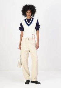 White cable-knit vest with blue accents over navy shirt, beige wide-leg trousers, and black shoes. Holding a small white bag.