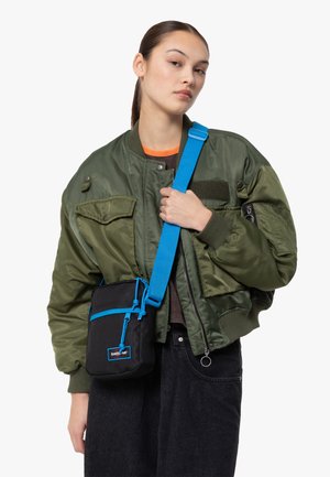 Sac noir avec des accents et une sangle bleus, de forme rectangulaire, en tissu texturé. Le modèle porte une veste bomber verte et un jean sombre.