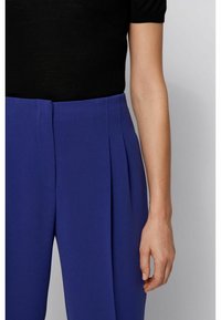 Un pantalon violet sur mesure avec des plis sur le devant, une texture lisse et une taille ajustée. Associé à un haut noir ajusté.