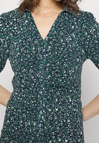 Femme portant une robe à zip florale verte et marine avec un col, les mains sur les hanches, sur un fond uni.