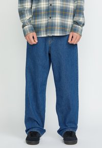 Jeans de mezclilla azul con un corte holgado, con cierre de botón y cremallera, combinados con una camisa de franela a cuadros en tonos gris, blanco y amarillo.