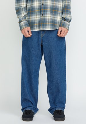 Blue denim jeans with a loose fit, featuring a button and zipper closure, worn with a plaid flannel shirt in gray, white, and yellow tones.
