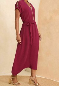 Viininpunainen mekko, jossa on lyhyet hihat, V-pääntie ja nauhavyötärö. Siinä on kultaiset yksityiskohdat ja joustava, nilkkapitkä muoto.