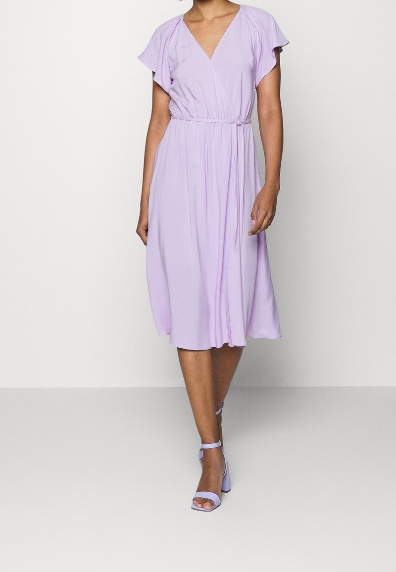 Robe lavande longueur genoux avec manches courtes volantées, col en V et taille élastique ; associée à des sandales à talons assorties à brides.