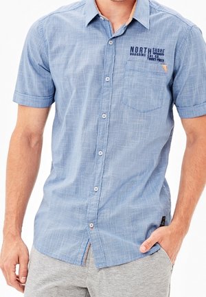 Lichtblauw overhemd met korte mouwen en knopen, met een borstzak en de tekst "NORTH SHORE", gedragen door een man met één hand in de zak van zijn grijze broek.