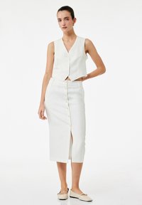 Witte cropped top met een V-hals en knopen aan de voorkant, gecombineerd met een witte knielange rok met een split aan de voorkant en een knoopsluiting.