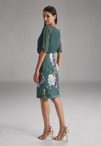 Robe florale verte avec des manches transparentes, de longueur genou, ornée de grandes fleurs roses et blanches. Associée à des sandales à talons hauts dorées.
