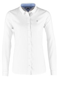 Camisa blanca de manga larga con cuello abotonado, interior del cuello en azul claro y pequeño logo en el lado izquierdo del pecho. Tela suave, diseño ajustado.