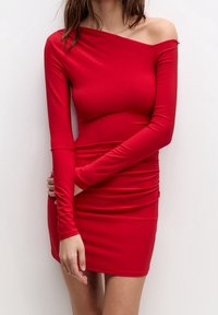 Robe moulante rouge à manches longues, design asymétrique avec une épaule dénudée. Fabriquée en matière stretch avec une texture froncée le long de la taille.