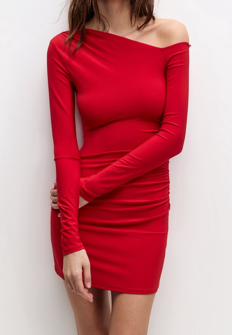 Robe moulante rouge à manches longues, design asymétrique avec une épaule dénudée. Fabriquée en matière stretch avec une texture froncée le long de la taille.