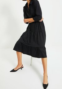 Abito nero a camicia con maniche a tre quarti, design a strati e vita legata, abbinato a tacchi a punta nera.