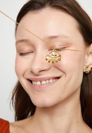 Collar con colgante de ojo dorado con detalles intrincados en una cadena delgada, llevado cerca del ojo. La modelo tiene un maquillaje ligero y una sonrisa suave.