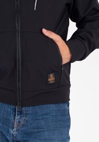 Giacca nera con zip, dalla texture liscia, con una tasca laterale e una toppa logo marrone. Accenti grigi con cordini sulla cappuccio.