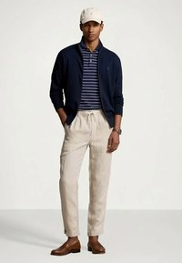 Marineblå glidelåssvegg over en marineblå og hvitstripet polo, kombinert med beige linbukser og brune skinnsko. Hvit caps på hodet.