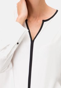 Blouse blanche avec des bordures noires, présentant un décolleté en V, des manches longues et un détail de bouton au niveau des poignets. Texture lisse.