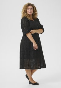 Robe noire en tissu à œillets avec motif chevron, manches bouffantes trois-quarts et fermeture à boutons. Assortie avec des chaussures plates noires.