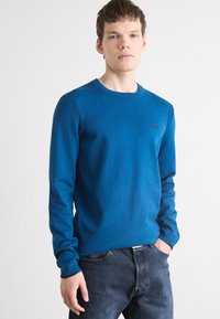 Pulover tricotat albastru cu mânecă lungă, cu accente cu nervuri la umeri și manșete. Prezintă un mic logo pe piept. Asociat cu blugi denim închis.