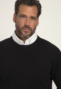 Schwarzer Strickpullover mit rundem Halsausschnitt, getragen über einem weißen Hemd mit Kragen. Der Stoff scheint weich zu sein und hat eine strukturierte Oberfläche.