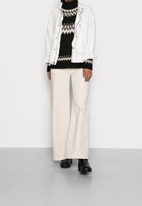 Weißer Blazer, schwarzer gemusterter Pullover, lockere beigefarbene Hose und schwarze klobige Stiefeletten. Hervorstechende Farben und unterschiedliche Texturen.