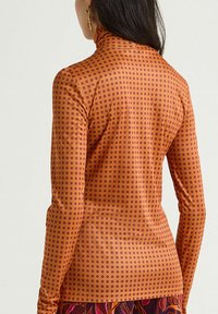 Haut à col roulé à manches longues orange avec un motif à pois violet. Texture lisse avec une silhouette ajustée et un col montant.