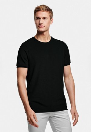 T-shirt noir à manches courtes en tissu lisse, avec un col rond et une coupe classique, associé à un pantalon de couleur claire.