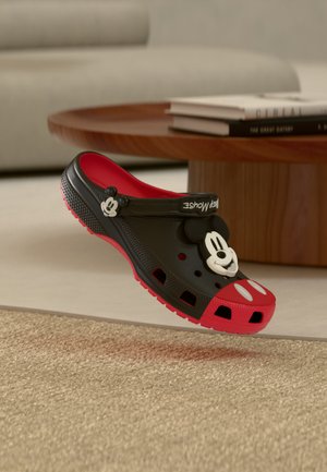Sabot noir et rouge avec décoration visage de Mickey Mouse à l'avant, bride réglable et trous de ventilation, placé près d'une table en bois sur un tapis.