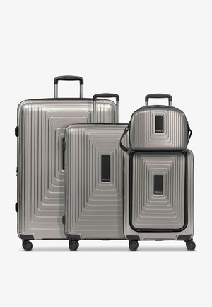 Quattro valigie rigide color argento con manici telescopici e ruote, di dimensioni variabili da grande a piccola, con etichette di marca nere al centro.