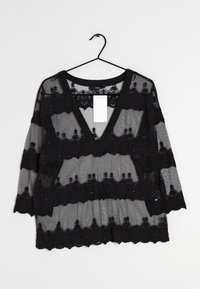 Blouse noire transparente avec un décolleté en V, mettant en avant des motifs de dentelle brodés le long des manches et du corps, et une bordure noire unie au niveau du col.