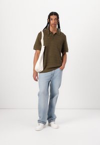 Polo shirt à manches courtes de couleur vert olive, texturé, associé à un jean bleu clair, large, et des baskets blanches, portant un sac en toile.