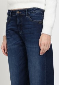 Donkerblauwe wide-leg jeans met een gladde denimtextuur, voorzien van een voorzakken en een metalen knoopdetail bij de taille.