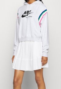 Jasnoszara przycięta bluza z kapturem Nike z czarnym logo, z różowo-niebiesko-zielonymi paskami na rękawach, zestawiona z białą spódnicą.