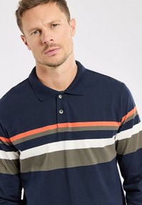 Polo shirt bleu marine avec des rayures horizontales oranges, vertes et blanches. Fabriqué en coton, avec un col et une patte de boutonnage à deux boutons.