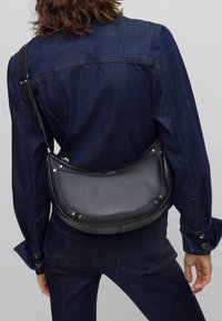 Borsa a spalla in pelle nera con forma arrotondata, superficie texturizzata e dettagli in metallo argentato, indossata sopra una giacca di denim scuro.