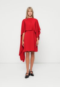 Rød kjole med en flytende kappeermedesign, laget av lett stoff. Kjolen har en A-linje form og er parret med svarte sko.