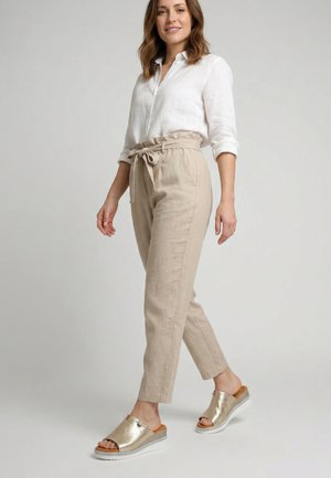 Kobieta nosząca beżowe spodnie z wysokim stanem w stylu paperbag z wiązanym paskiem, białą bluzkę z podwiniętymi rękawami oraz metaliczne złote sandały na platformie wsuwane.