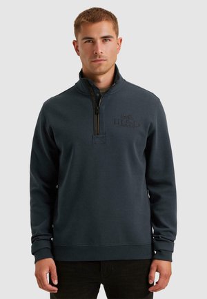 Young man wearing a dark blue quarter-zip sweatshirt with "PME Legend" embroidered on the chest, standing against a plain background.