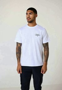 Man with tattoos on both arms wearing a white t-shirt with small black text on chest and black pants, standing against plain background.
