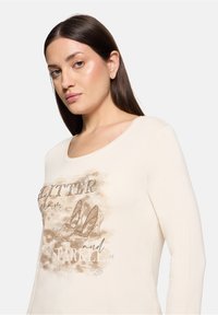 Beiges Langarmshirt mit rundem Ausschnitt, verziert mit einem Design aus High Heels und Text in schimmernden Strasssteinen und hellbraunen Akzenten.