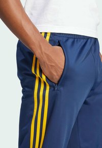 Marina atletiska byxor med gula ränder längs sidorna, elastisk midja och fickor med dragkedja. Mjuk, slät textur.