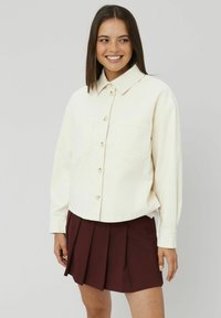 Chemise en velours côtelé crème avec un devant à boutons, un col et deux poches poitrine, associée à une jupe plissée bordeaux.