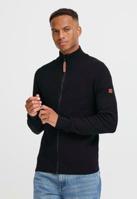 Suéter negro con cremallera y cuello alto, puños acanalados y una etiqueta de cuero en la manga izquierda, hecho de una tela suave y texturizada.