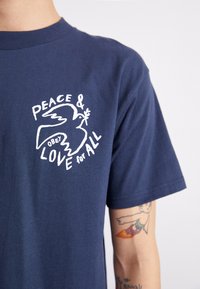 T-shirt de algodão azul-marinho com uma gráfica branca de uma pomba com um ramo e o texto: "PAZ & OBEDECE AMOR a TODOS." Mangas curtas.