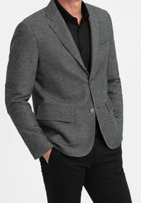 Homme portant un blazer gris texturé sur une chemise noire à boutons, les mains dans les poches de son pantalon noir, sur un fond uni.