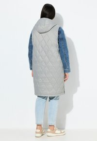 Gesteppte graue Wattierte Weste mit einem Jeansärmel und Kapuze, mit einem mittellangen Design und strukturierter Stoff. Wird über hellblauen Jeans getragen.